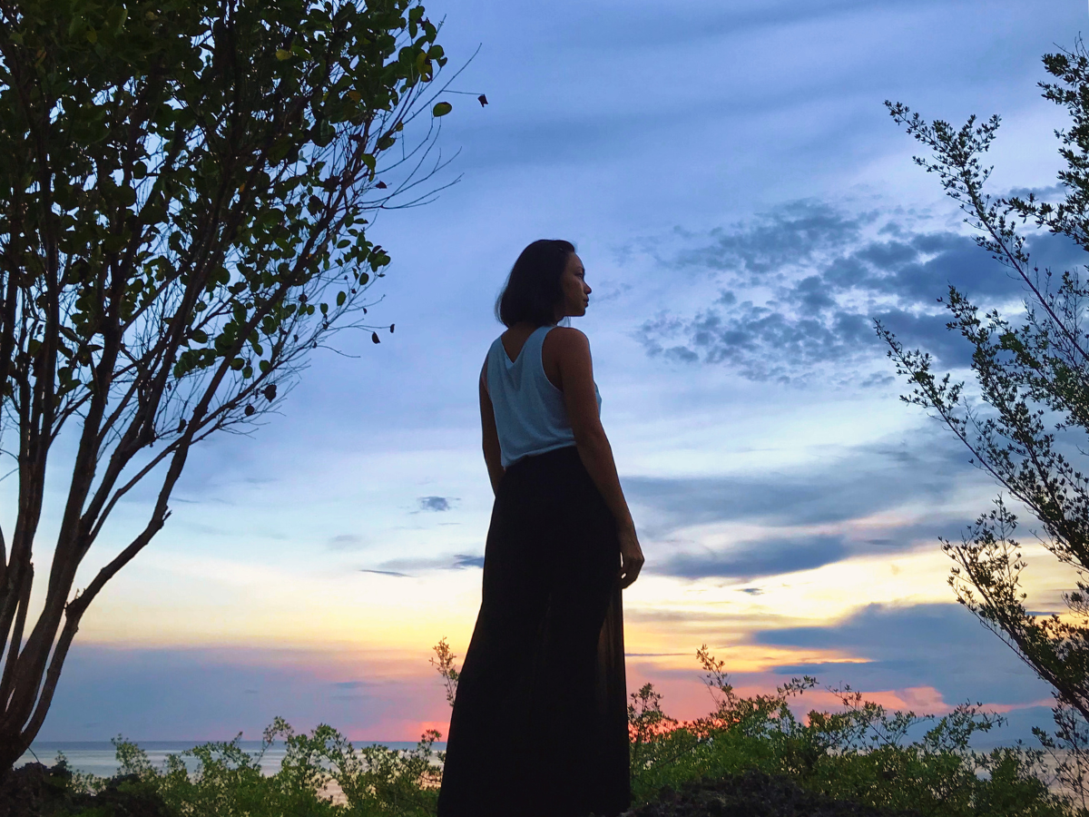 A mom learning to care for herself as a whole and thrive in motherhood, standing on a cliff watching the sunset.