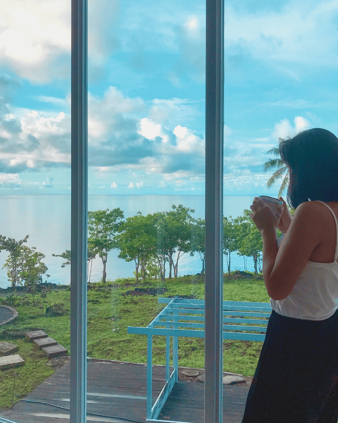 Lady having a serene and peaceful morning, drinking coffee looking over the glass window with a beautiful beach view and nature