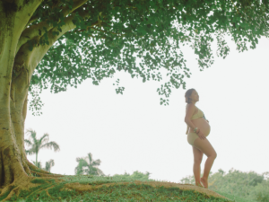 A pregnant woman stands thoughtfully under a large tree, cradling her baby bump as she reflects on her birth story.