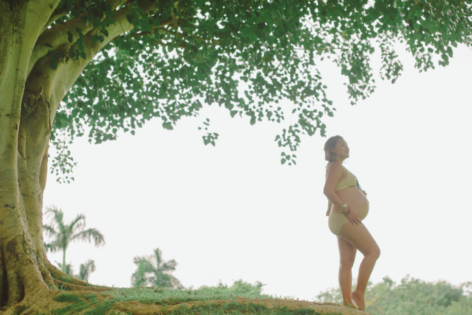 A pregnant woman stands thoughtfully under a large tree, cradling her baby bump as she reflects on her birth story.