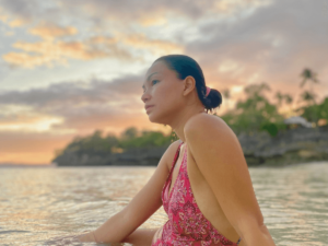 A mother sitting in the sea, gazing thoughtfully toward the horizon, reflecting on her life-changing Self-Care Journey with a beautiful backdrop of orange skies and a vibrant sunset.