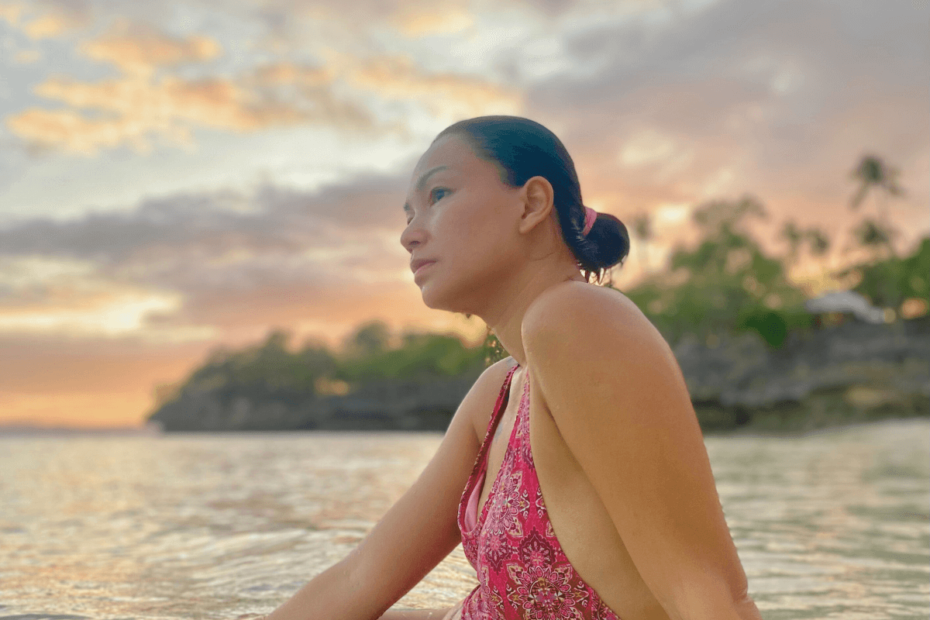 A mother sitting in the sea, gazing toward the horizon, reflecting thoughtfully on her Self-Care Journey, framed by orange skies and a vibrant sunset.