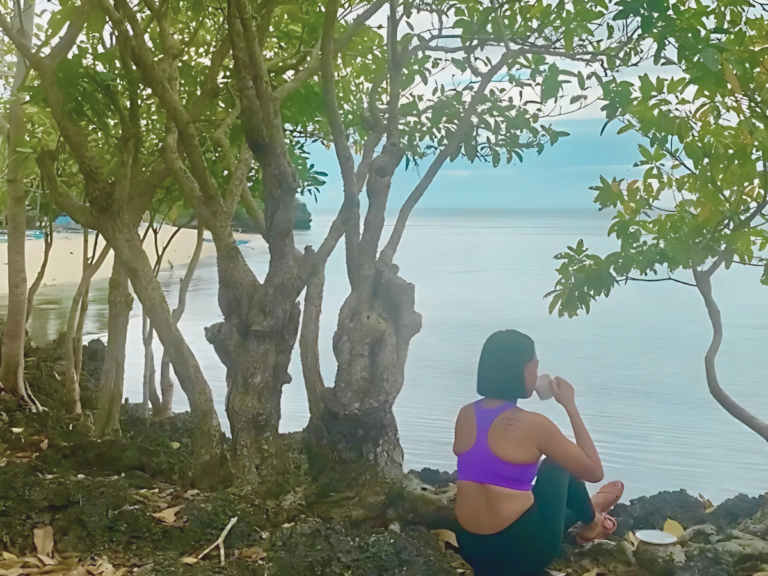 Woman sitting by a cliff, gazing out at the ocean while drinking her morning coffee, embracing the calmness of the moment and practicing mindfulness.