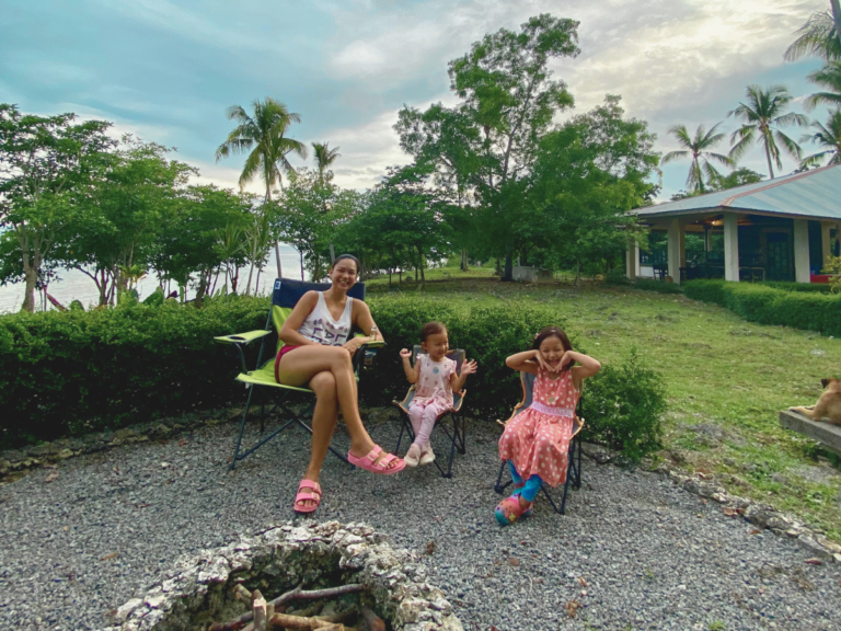 Mother and her two daughters sitting on picnic chairs, smiling and looking at the camera, enjoying a peaceful moment before the sun sets to start a bonfire—perfect example of listening more and connecting as a family.
