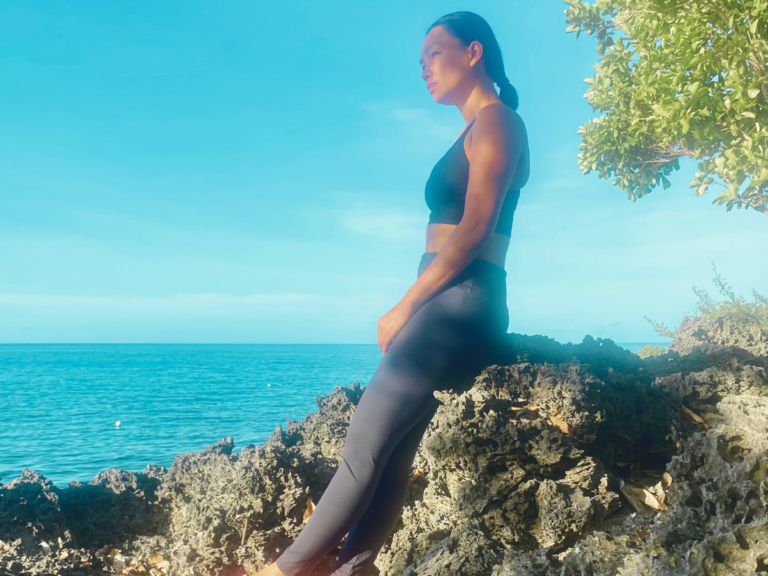 a mother in her work-out clothes leaning on a rock enjoys the view of the ocean and the morning sun as part of her simple morning habits