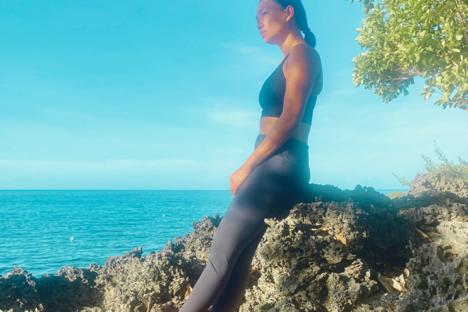 a mother in her work-out clothes leaning on a rock enjoys the view of the ocean and the morning sun as part of her simple morning habits