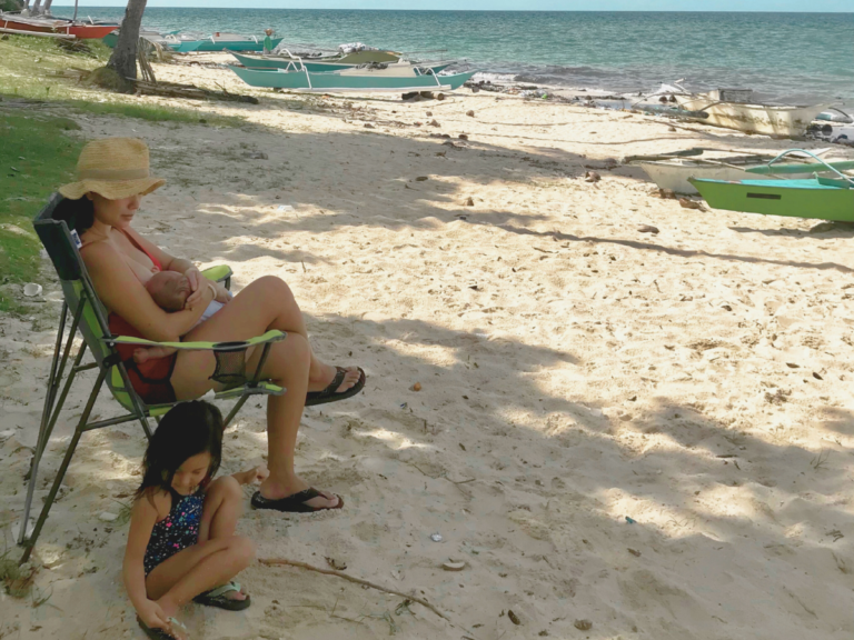 A mother sitting on a beach chair by the ocean, breastfeeding her newborn while her toddler plays in the sand, reflecting on how to prepare for motherhood.