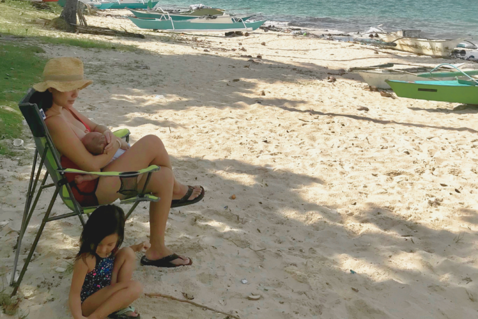 A mother sitting on a beach chair by the ocean, breastfeeding her newborn while her toddler plays in the sand, reflecting on how to prepare for motherhood.