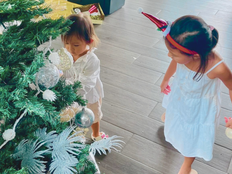 Two little girls decorating the Christmas tree in preparation for Christmas on the Island celebrations. The eldest wears a white dress and Christmas hat headband, while the toddler wears a white blouse.
