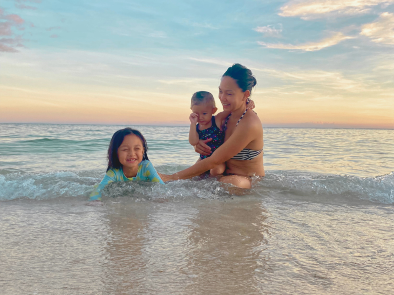 A mom enjoying the beach with her two daughters, carrying her toddler while laughing with her eldest daughter sitting on the sand, reflecting on how she can become a great mom.
