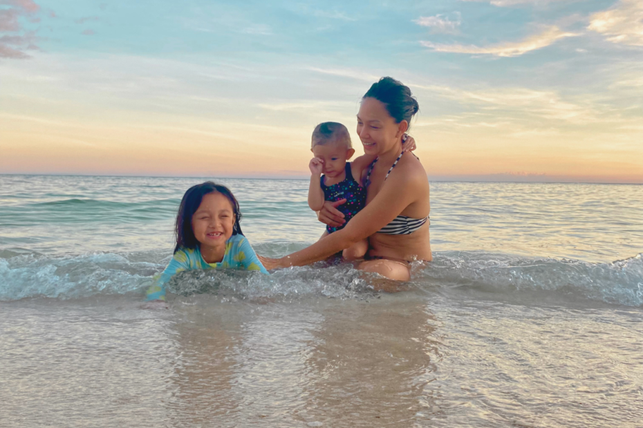 A mom enjoying the beach with her two daughters, carrying her toddler while laughing with her eldest daughter sitting on the sand, reflecting on how she can become a great mom.