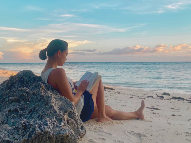 A mother leaning against a rock on the beach by sunset, reading a book, embracing self-care and setting healthy boundaries to prioritize her well-being and energy for her family.