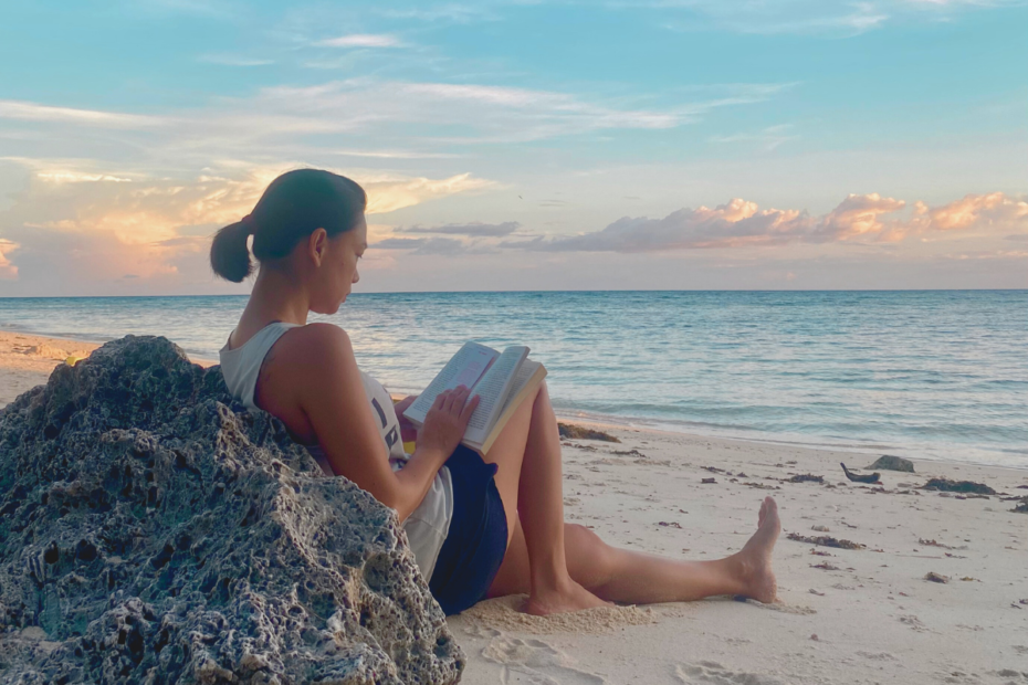 A mother leaning against a rock on the beach by sunset, reading a book, embracing self-care and setting healthy boundaries to prioritize her well-being and energy for her family.