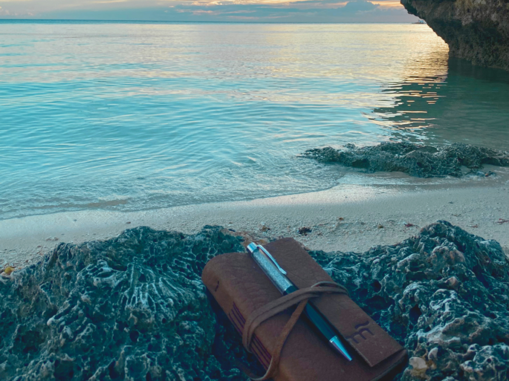A notebook and pen resting on a rock by the beach, illuminated by a stunning sunset, a serene setting to crush your goals.