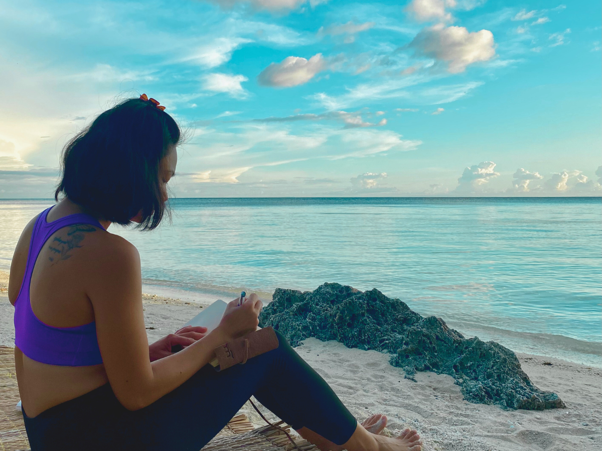 A mom sitting on the beach at sunset, writing in her notebook, reflecting on how to crush your goals with the calming ocean view as inspiration.