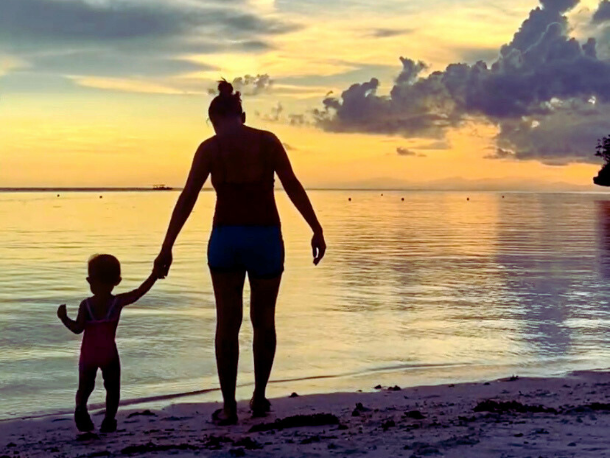 A mother holding her todller's hand as they walk towards the water on the beach during sunset, navigating postpartum.
