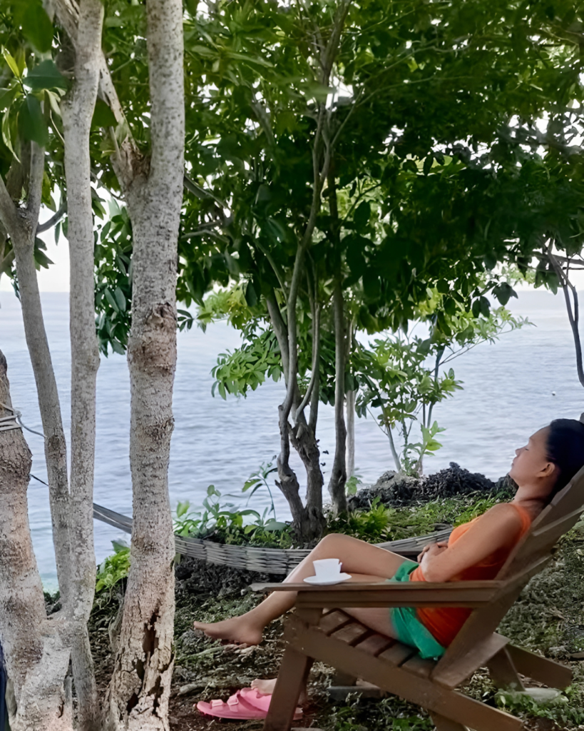 An overstimulated mom sits by a hammock, eyes closed, savoring a quiet moment with her coffee, finding relief from the chaos of motherhood.