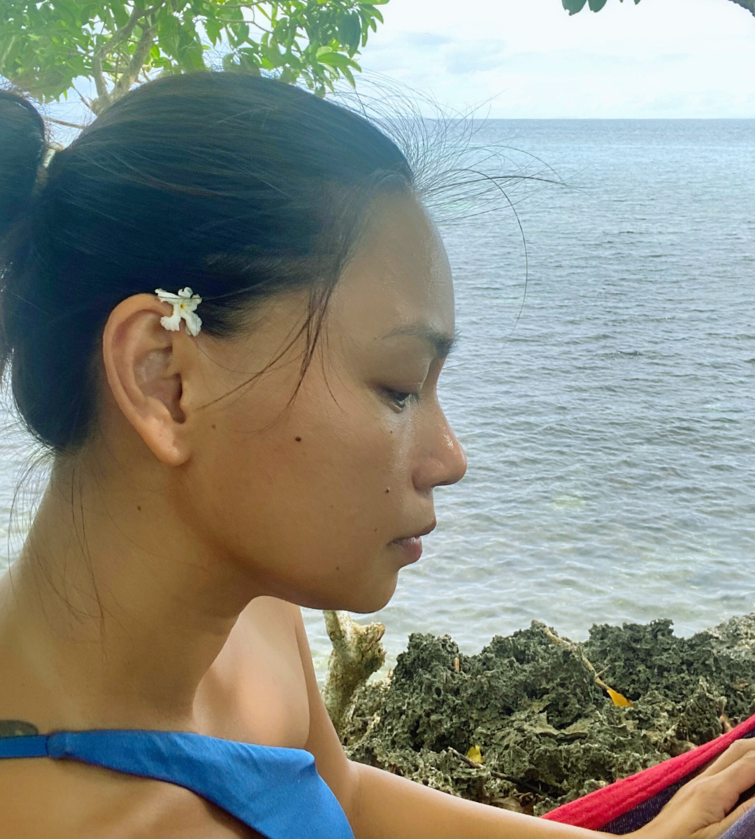 A woman with a small white flower on her right ear, sitting on a hammock on a cliff with a view of the ocean, enjoying her alone time practicing self-care for moms.
