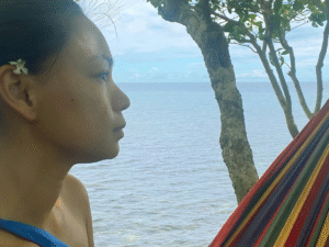 A woman thinking she's more than just a mom with a small white flower on her right ear, sitting on a hammock on a cliff with a view of the ocean.