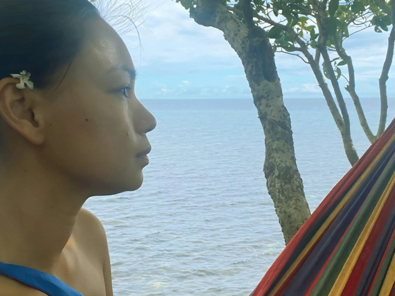 A woman with a small white flower on her right ear, sitting on a hammock on a cliff with a view of the ocean, wondering if she can love her children and still want to be more than just a mom