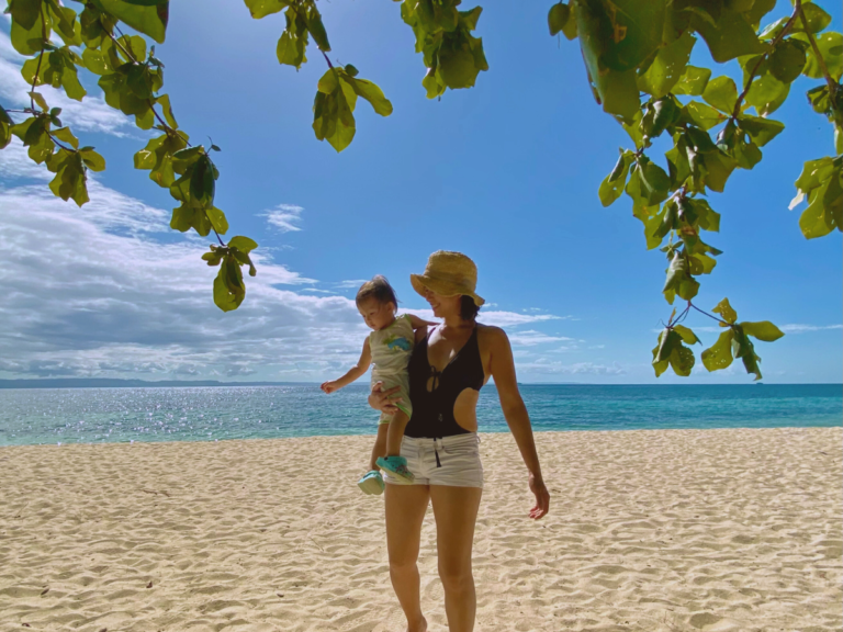 A mom quietly wondering why the transition to motherhood is so hard, carrying her baby on a white sand beach on a birght, sunny day with a beautiful view of the sparkling ocean and the blue sky.