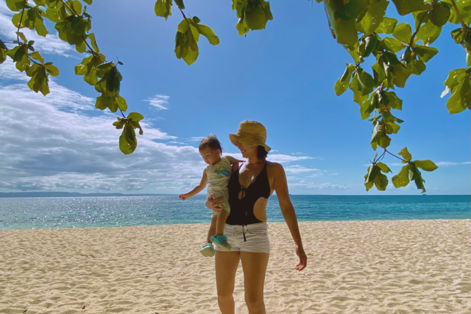 A mom quietly wondering why the transition to motherhood is so hard, carrying her baby on a white sand beach on a birght, sunny day with a beautiful view of the sparkling ocean and the blue sky.