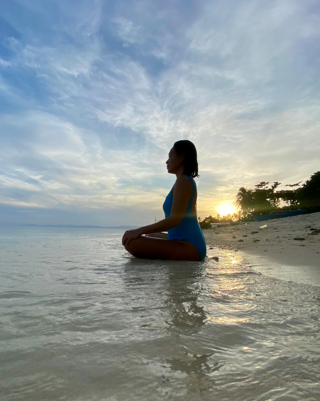 An island mom sitting on the beach with a view of the sunset, meditating as part of her self-care.