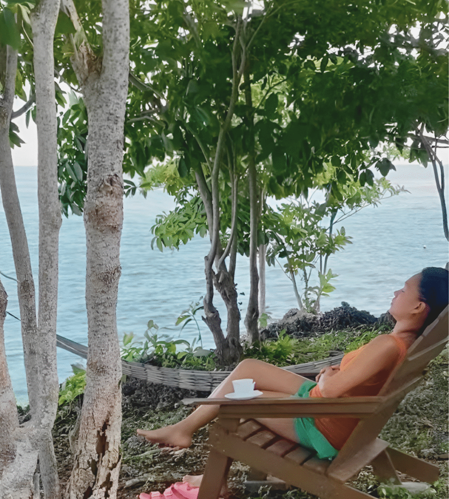 An overstimulated mom sits by a hammock, eyes closed, savoring a quiet moment with her coffee, finding relief from the chaos of motherhood.