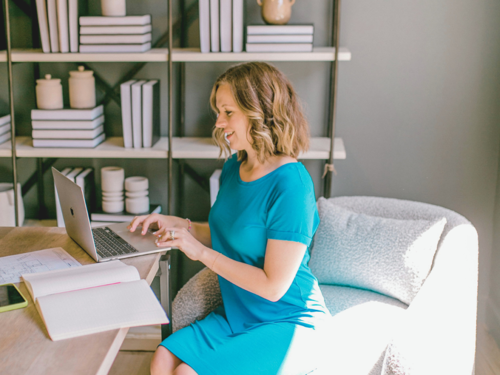 A mom wearing a blue dress sitting on a desk while happily working on her laptop
