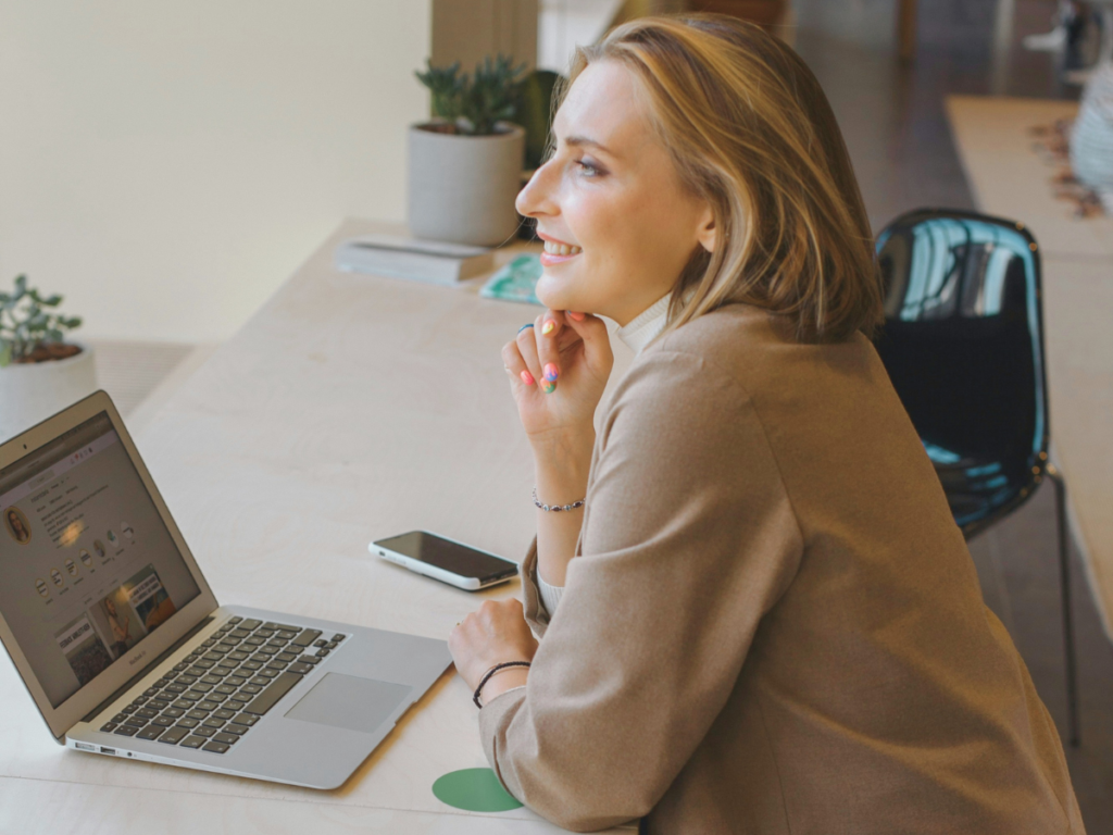 A smiling mom in a brown coat, sitting on a desk, working on her Mac